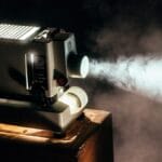 A vintage film projector beams light through smoke in a dimly lit room, sitting on a wooden box.