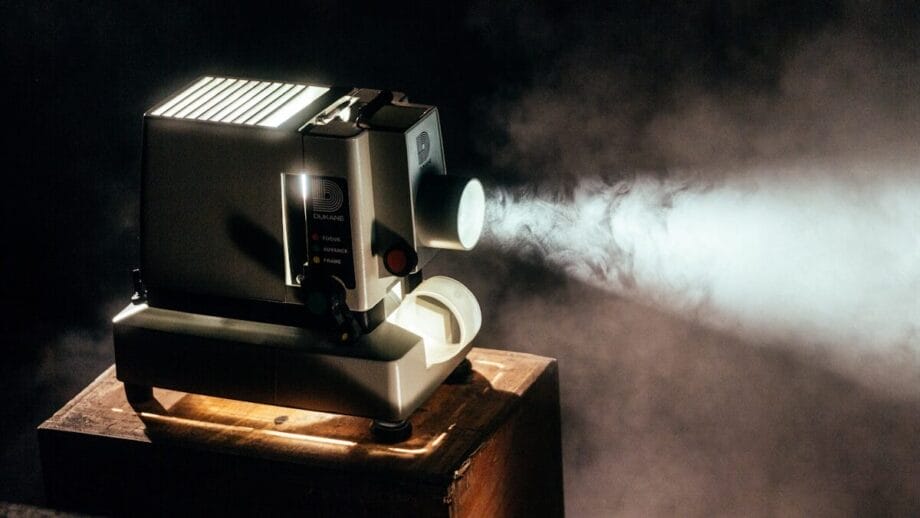 A vintage film projector beams light through smoke in a dimly lit room, sitting on a wooden box.