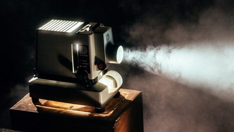 A vintage film projector beams light through smoke in a dimly lit room, sitting on a wooden box.