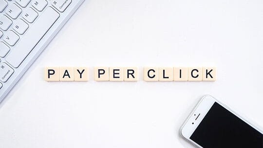 Letter tiles spelling Pay Per Click on a white surface, with a keyboard and smartphone nearby.