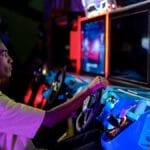 Teen playing a racing video game at an arcade. He sits in a blue seat, focused on the screen ahead, holding the steering wheel.