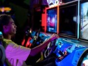 Teen playing a racing video game at an arcade. He sits in a blue seat, focused on the screen ahead, holding the steering wheel.