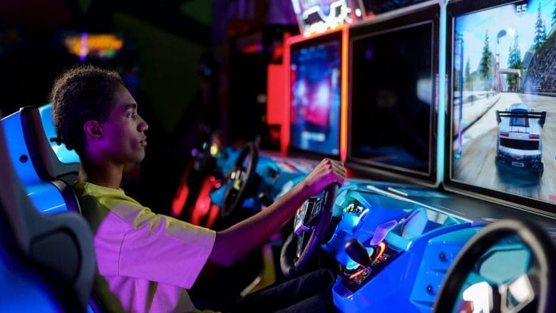 Teen playing a racing video game at an arcade. He sits in a blue seat, focused on the screen ahead, holding the steering wheel.