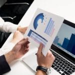 Two people review printed charts and a bar graph displayed on a laptop screen at a white desk in an office setting.