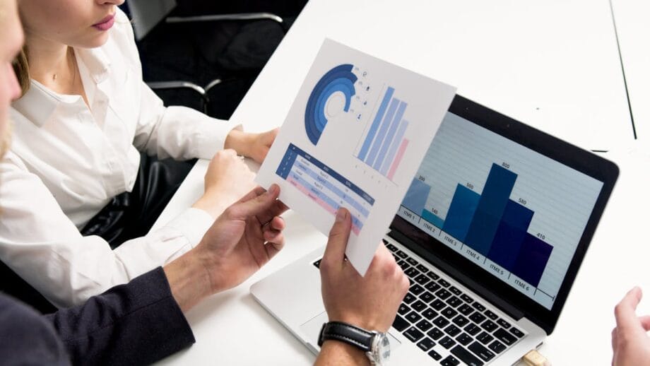 Two people review printed charts and a bar graph displayed on a laptop screen at a white desk in an office setting.