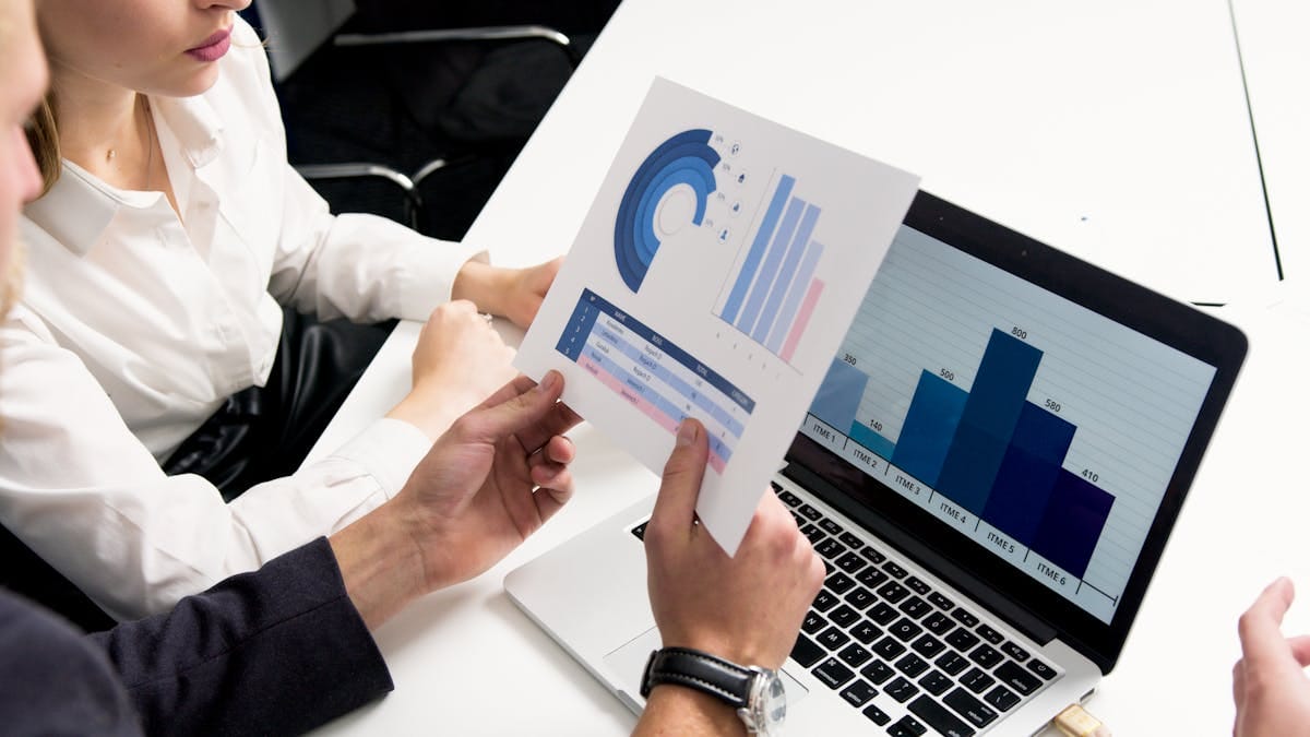 Two people review printed charts and a bar graph displayed on a laptop screen at a white desk in an office setting.