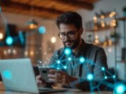 Man with glasses using smartphone and laptop in a warmly lit room, surrounded by digital network graphics.