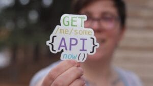 Person holding a sticker that reads GET /me/some {API} now with a blurred background.