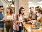 Five people collaborate around a wooden table with laptops and tablets in a modern office setting.