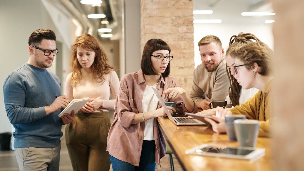 Five people collaborate around a wooden table with laptops and tablets in a modern office setting.