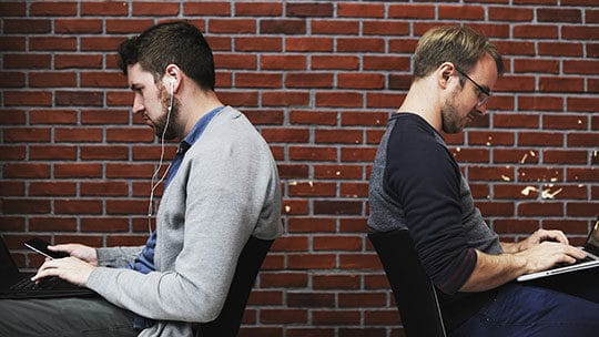 Two men sit back-to-back against a brick wall, using laptops; one is wearing earphones.