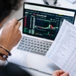 Person holding a printed spreadsheet points at a laptop screen displaying data and charts in an office setting.