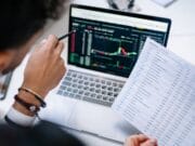 Person holding a printed spreadsheet points at a laptop screen displaying data and charts in an office setting.
