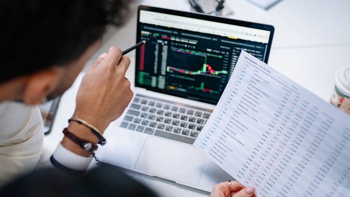 Person holding a printed spreadsheet points at a laptop screen displaying data and charts in an office setting.