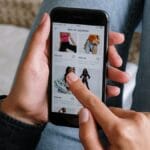 A person browses clothing items on a smartphone while sitting with legs crossed on a bed.