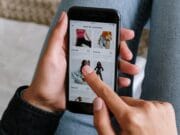 A person browses clothing items on a smartphone while sitting with legs crossed on a bed.