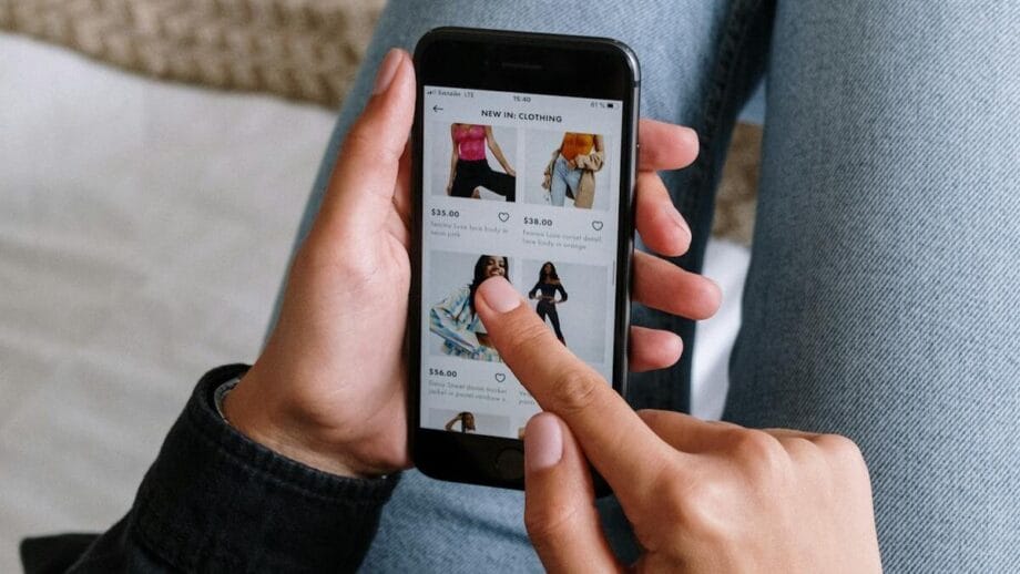 A person browses clothing items on a smartphone while sitting with legs crossed on a bed.