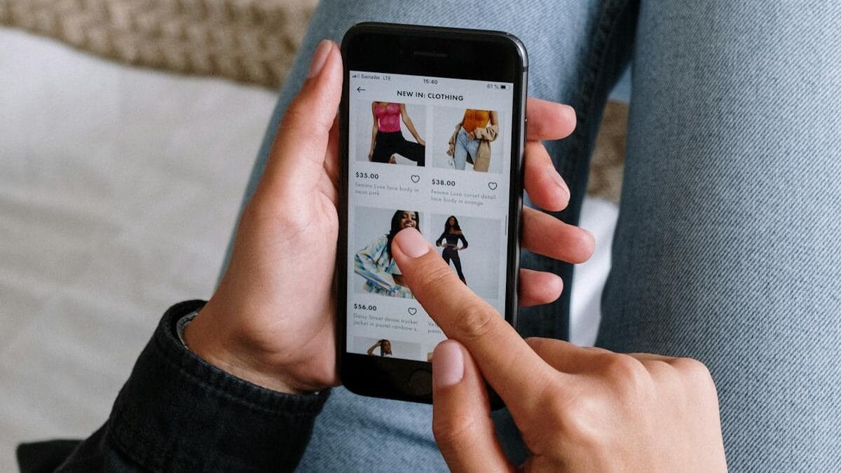 A person browses clothing items on a smartphone while sitting with legs crossed on a bed.
