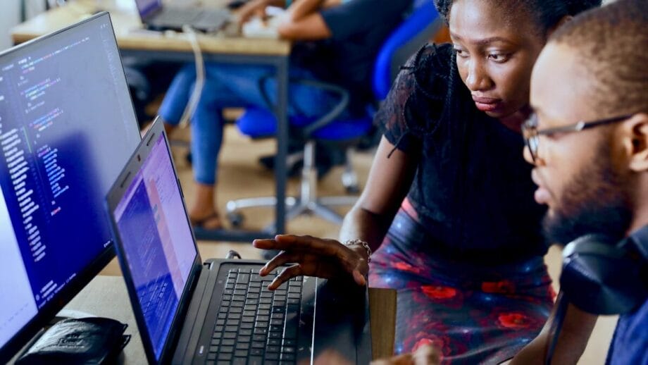 Two people collaborate at a desk with laptops, reviewing code on the screen, while others work in the background.