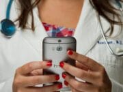 A person in a white lab coat with a stethoscope is holding and using an HTC smartphone.