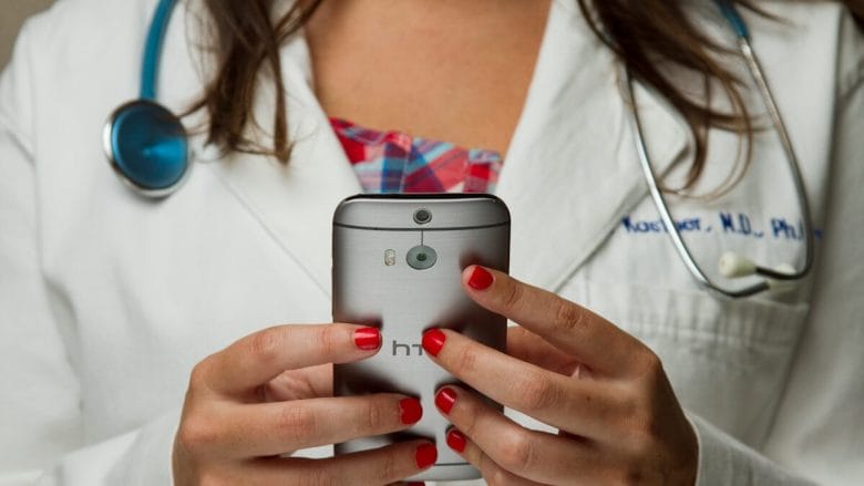 A person in a white lab coat with a stethoscope is holding and using an HTC smartphone.