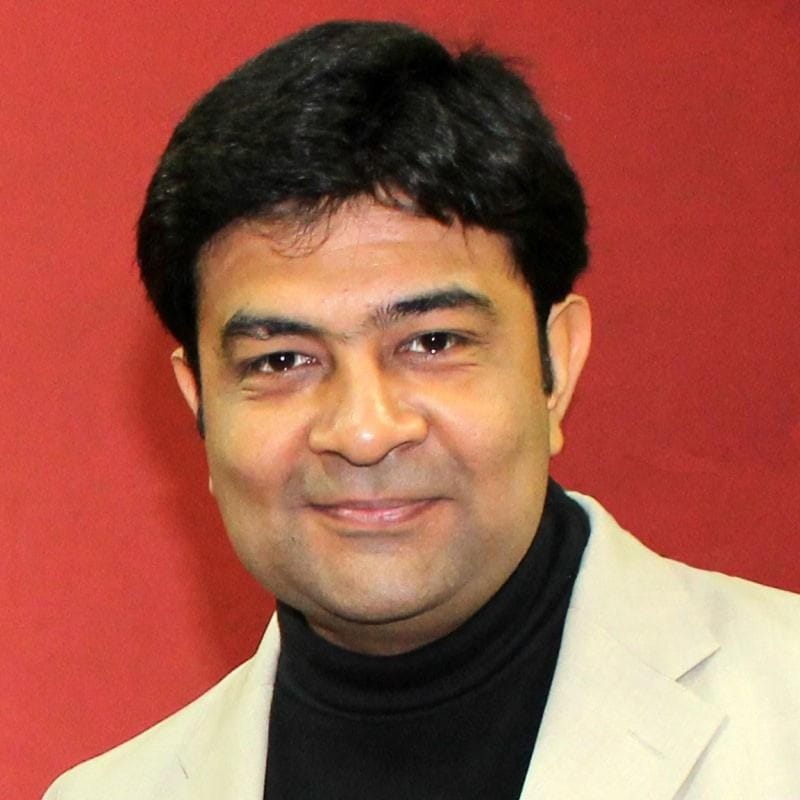 Souvik Banerjee with short black hair wearing a beige blazer and black turtleneck stands in front of a plain red background. Souvik Banerjee with short black hair wearing a beige blazer and black turtleneck stands in front of a plain red background.