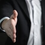 A person in a suit extends their right hand forward, as if offering a handshake.