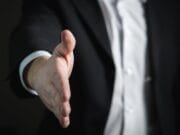 A person in a suit extends their right hand forward, as if offering a handshake.