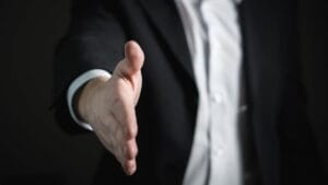 A person in a suit extends their right hand forward, as if offering a handshake.