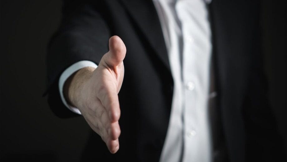 A person in a suit extends their right hand forward, as if offering a handshake.