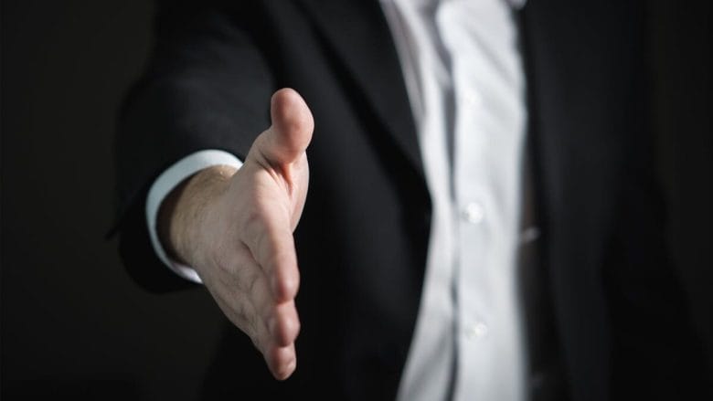 A person in a suit extends their right hand forward, as if offering a handshake.
