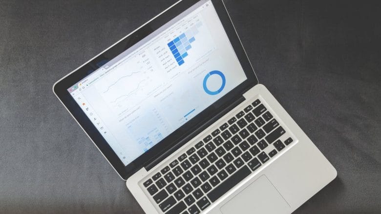 Open laptop displaying data charts and graphs on the screen, placed on a dark surface with a wooden floor visible.
