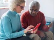 Two older adults sit on a couch; one holds a smartphone and the other holds a credit card, appearing to make an online transaction.