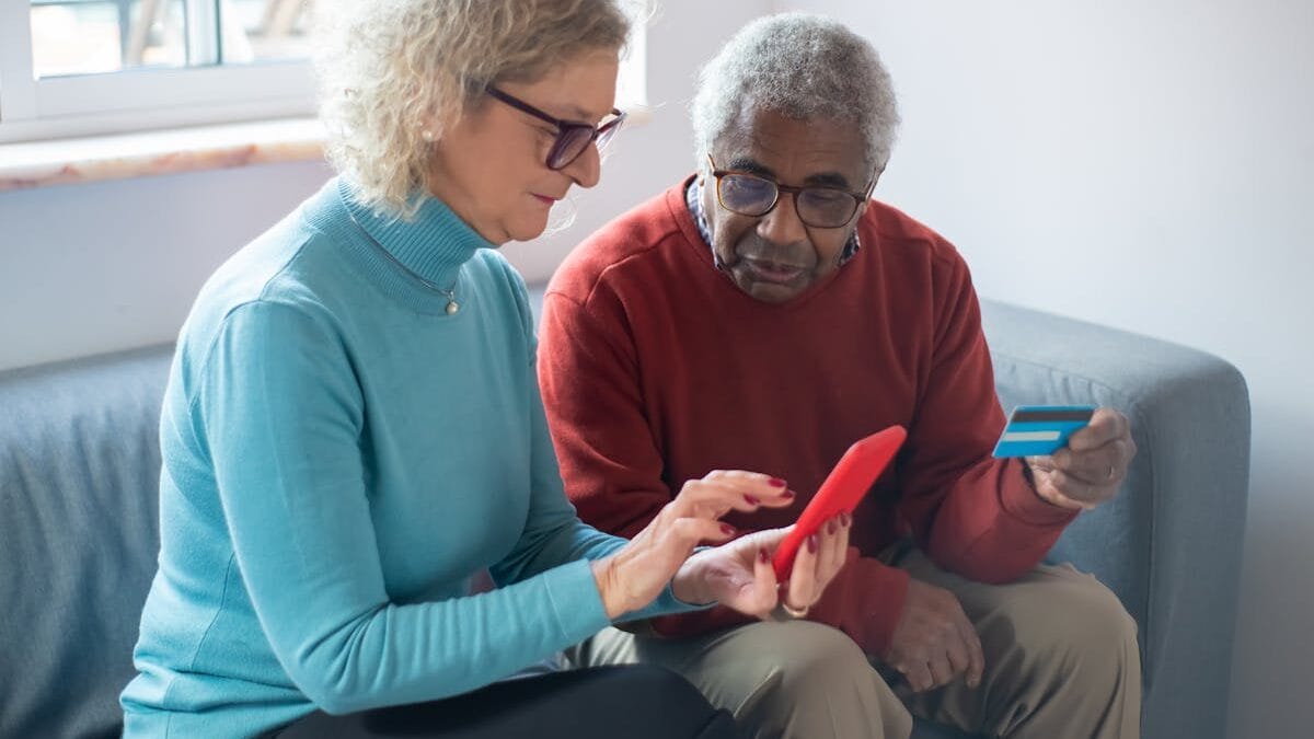 Two older adults sit on a couch; one holds a smartphone and the other holds a credit card, appearing to make an online transaction.
