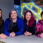Four women stand together at a counter, smiling, with a wooden wall and a yellow geometric sign in the background.