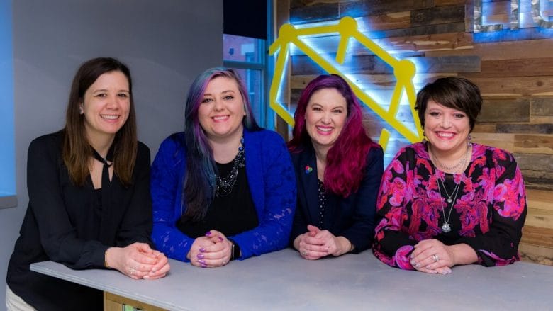 Four women stand together at a counter, smiling, with a wooden wall and a yellow geometric sign in the background.