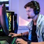 A young man wearing headphones and a microphone sits at a desk, gaming on a computer with a backlit keyboard and large monitor.