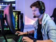 A young man wearing headphones and a microphone sits at a desk, gaming on a computer with a backlit keyboard and large monitor.