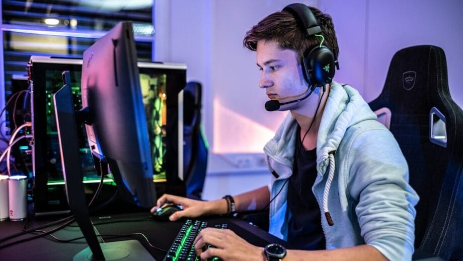 A young man wearing headphones and a microphone sits at a desk, gaming on a computer with a backlit keyboard and large monitor.