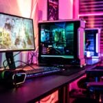 A gaming setup with a desktop PC, monitor, keyboard, mouse, headphones, and LED lighting in a room with blinds.