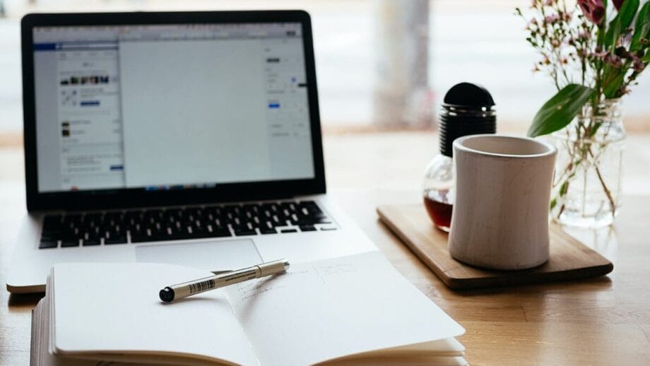 Open laptop, notebook with pen, coffee mug, and flowers on a wooden desk, suggesting a workspace or study area.
