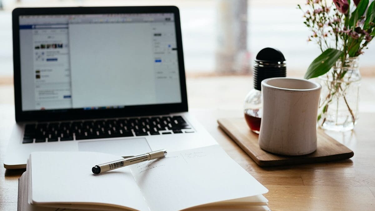 Open laptop, notebook with pen, coffee mug, and flowers on a wooden desk, suggesting a workspace or study area.
