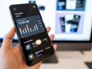 A hand holds a smartphone displaying a financial app with spending data; a computer screen is blurred in the background.
