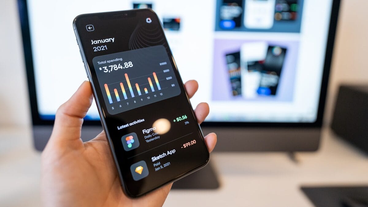 A hand holds a smartphone displaying a financial app with spending data; a computer screen is blurred in the background.