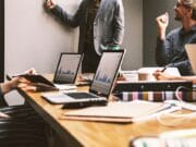 Four people in a meeting room with laptops open, one person standing and presenting at a whiteboard, others seated and listening.