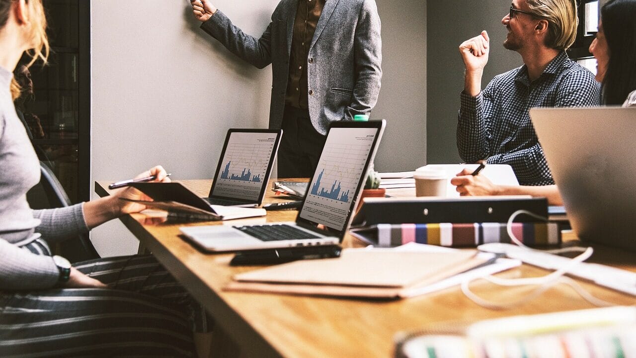 Four people in a meeting room with laptops open, one person standing and presenting at a whiteboard, others seated and listening.