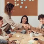 Six people sit at a table in a meeting room, with two of them shaking hands and a corkboard with sticky notes in the background.