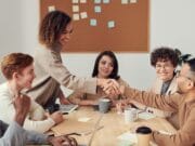 Six people sit at a table in a meeting room, with two of them shaking hands and a corkboard with sticky notes in the background.