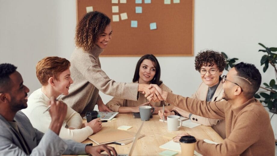 Six people sit at a table in a meeting room, with two of them shaking hands and a corkboard with sticky notes in the background.