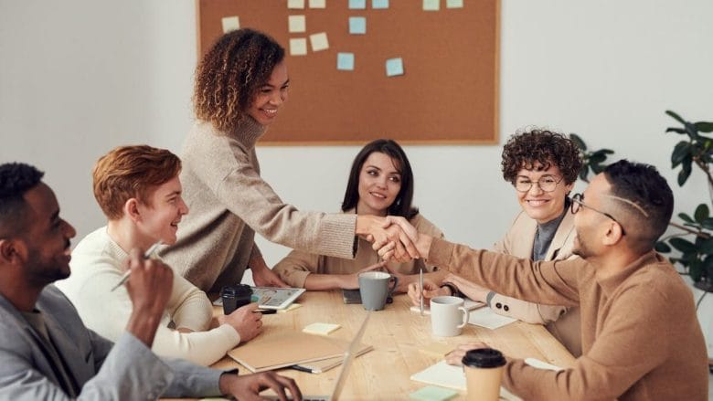Six people sit at a table in a meeting room, with two of them shaking hands and a corkboard with sticky notes in the background.